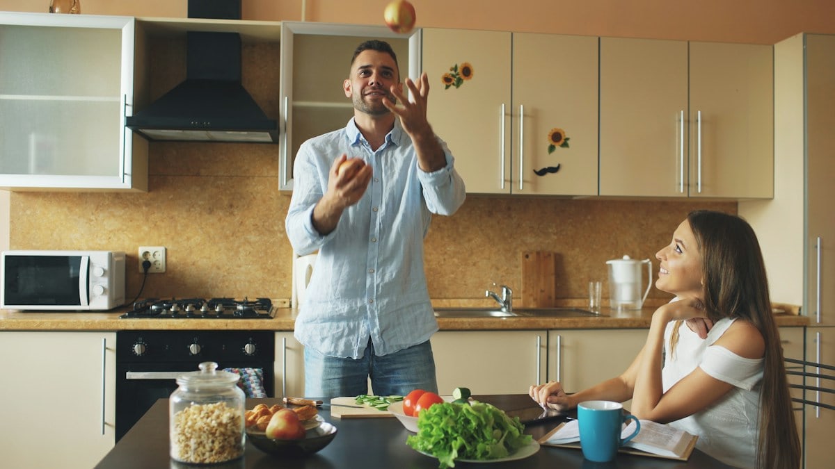 Attractive loving couple having fun in the kitchen. Handsome man juggle with apples to impress his girlfriend at home