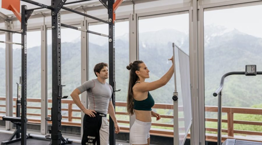 A gym coach instructs on whiteboard beside fitness equipment with mountain view.